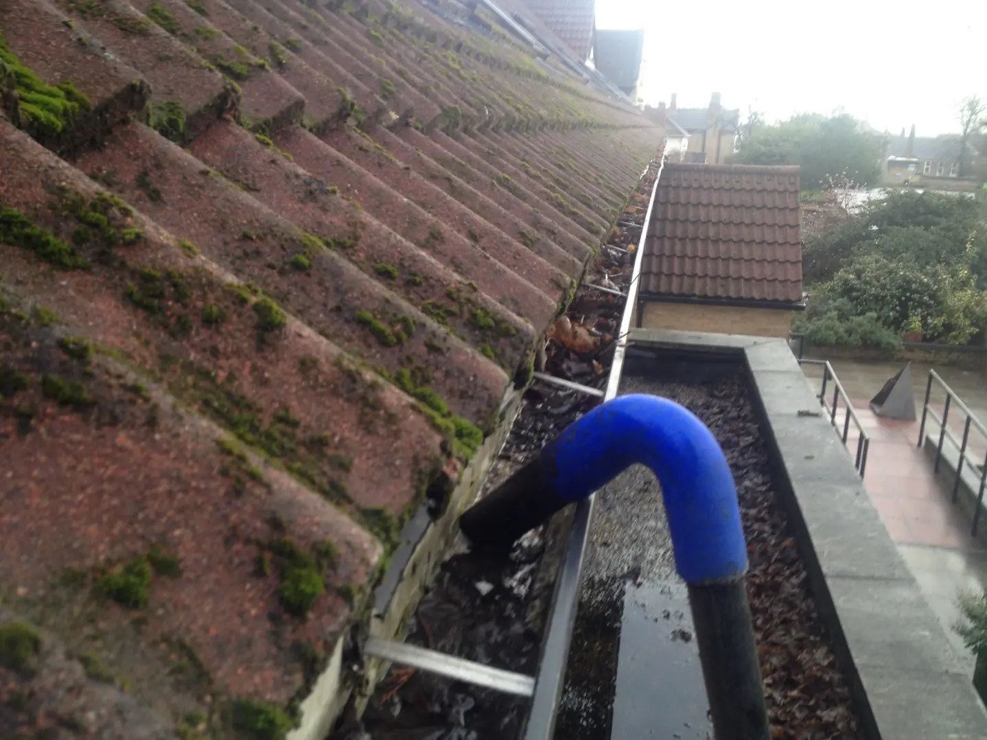 A close-up view of a gutter clogged with leaves, dirt, and debris. The gutter is visibly dirty, with water stains and signs of neglect. Overhanging leaves and twigs are blocking the water flow, leading to potential water damage and inefficiency.